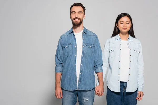 Stylish interracial couple in denim clothes and gumshoes holding hands while standing with closed eyes on grey — Stock Photo