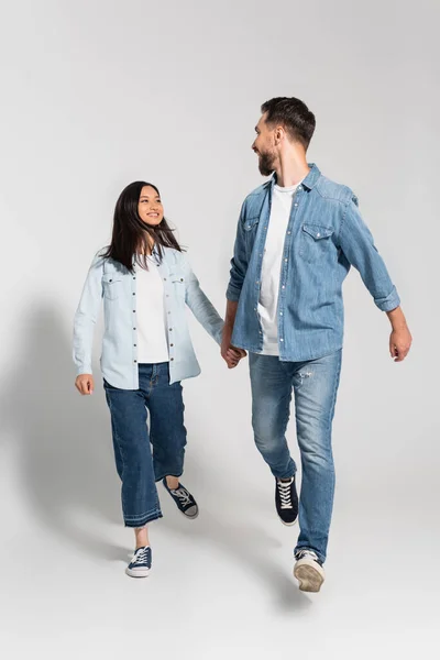 Elegante interracial casal no denim roupas segurando as mãos e olhando para o outro enquanto caminhando no cinza — Fotografia de Stock