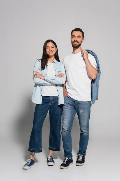 Longitud completa con de elegante pareja interracial en ropa de mezclilla y zapatos de goma mirando a la cámara en gris - foto de stock