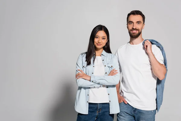 Mujer asiática elegante con brazos cruzados cerca de hombre barbudo con la mano en el bolsillo en gris - foto de stock