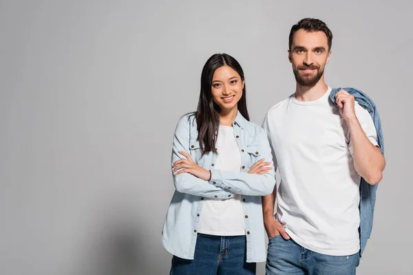 Young asian woman in denim shirt standing with crossed arms near boyfriend with hand in pocket on grey — Stock Photo