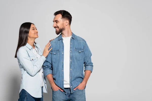 Young asian woman touching bearded boyfriend standing with hands in pockets on grey — Stock Photo