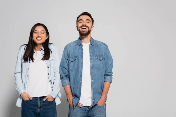 Young interracial couple in denim clothes holding hands in pockets while standing with closed eyes on grey — Stock Photo