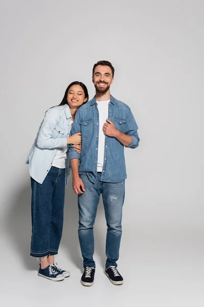 Full length view of stylish asian woman leaning on bearded boyfriend in denim clothes on grey — Fotografia de Stock