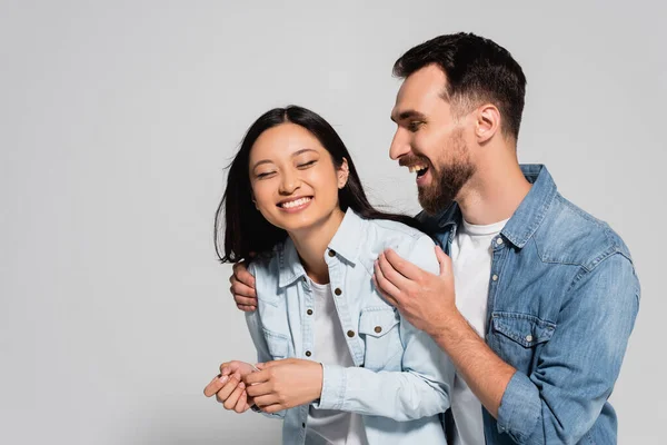 Excited man touching shoulders of asian girlfriend in denim shirt on grey — Stock Photo