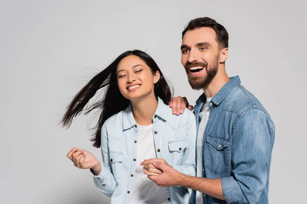 Eccitata, elegante coppia interrazziale in camicie di jeans tenendosi per mano sul grigio — Foto stock