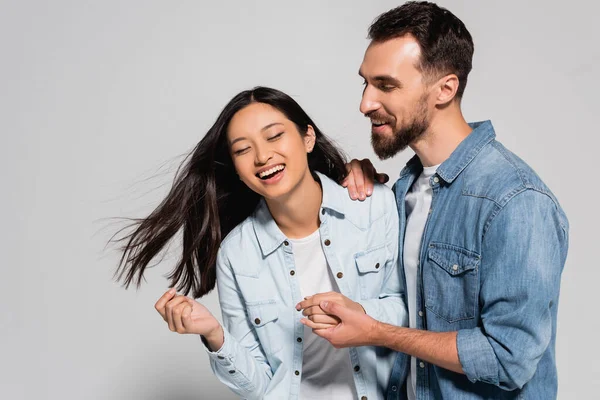 Bärtiger Mann hält Händchen mit stylischer asiatischer Freundin und lacht mit geschlossenen Augen auf grau — Stockfoto