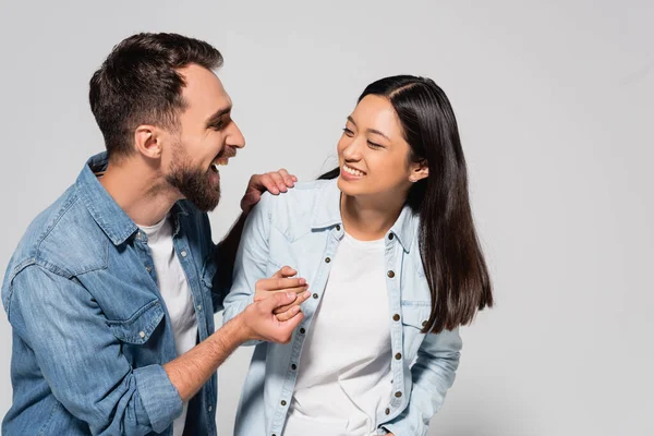 Excited man laughing while holding hands with asian girlfriend on grey — Stock Photo