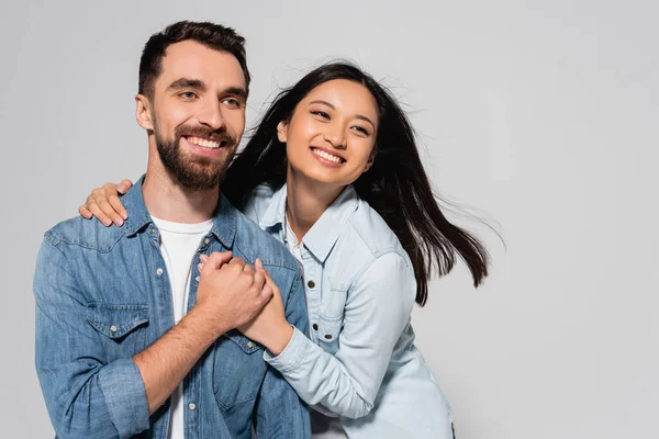 Stylish interracial couple in denim shirts holding hands and looking away isolated on grey — Stock Photo