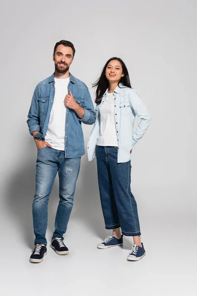 Повний вигляд молодої, стильної міжрасової пари в джинсовому одязі, дивлячись на камеру на сірому — стокове фото