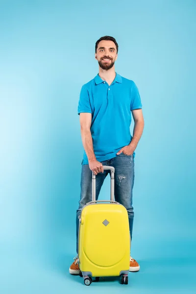 Young tourist in polo t-shirt and jeans holding hand in pocket while standing with yellow suitcase on blue — Stock Photo