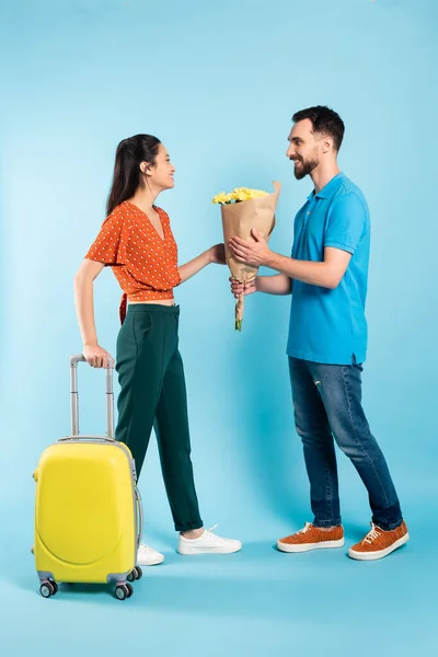 Hombre en polo camiseta y jeans presentando ramo a mujer asiática con maleta amarilla en azul - foto de stock
