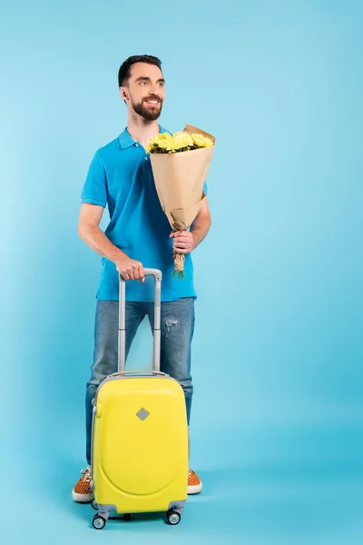 Joven turista en jeans y camiseta de polo sosteniendo ramo mientras está de pie con la maleta amarilla en azul - foto de stock