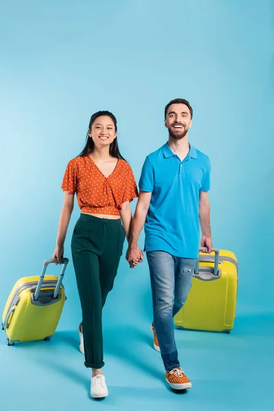 Visão de comprimento total de casal interracial de turistas de mãos dadas enquanto caminhava com malas em azul — Fotografia de Stock