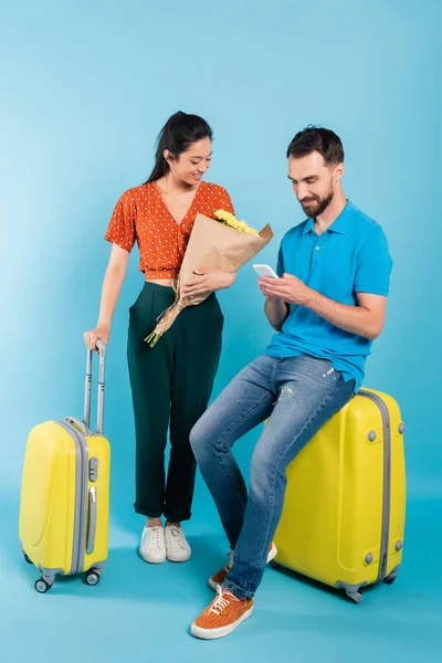 Asian woman in red blouse looking at bouquet near boyfriend using smartphone while sitting on suitcase on blue — Stock Photo