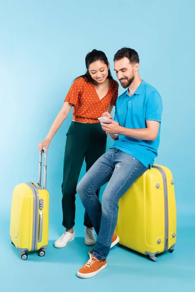 Joven hombre en polo camiseta usando teléfono inteligente mientras está sentado en la maleta cerca de mujer asiática en blusa roja en azul - foto de stock
