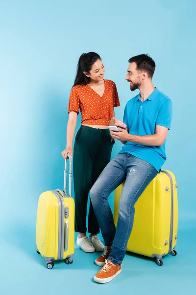 Asiatin in roter Bluse schaut Freund auf Koffer an und zeigt mit der Hand auf Smartphone auf blauem Grund — Stockfoto