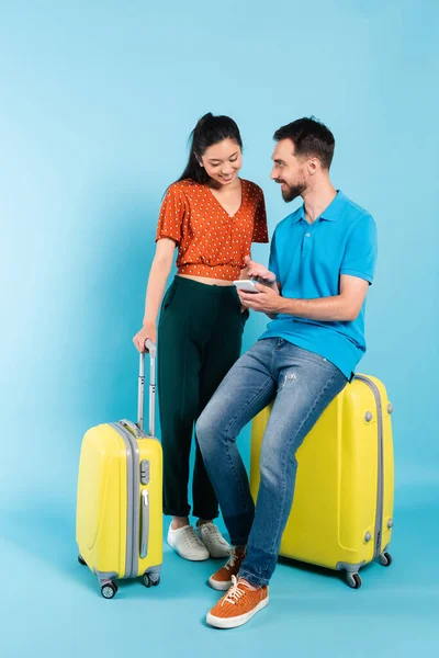 Bearded man in polo t-shirt pointing with hand at smartphone while sitting on suitcase near asian girlfriend on blue — Stock Photo