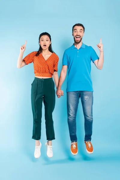 Excitada pareja asiática cogida de la mano y mostrando gesto de idea mientras levita sobre azul - foto de stock