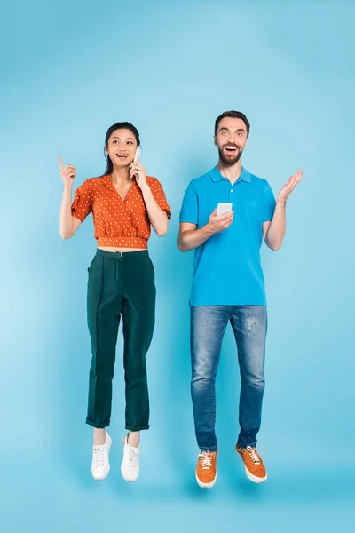 Morena asiática mujer hablando en smartphone y apuntando con el dedo cerca excitado hombre levitando sobre azul - foto de stock