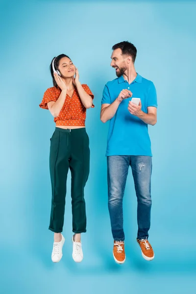 Excitado hombre apuntando con el dedo en el teléfono inteligente cerca complacido mujer asiática en los auriculares levitando en azul - foto de stock