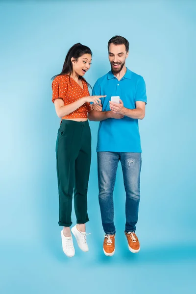 Excitada mujer asiática señalando con el dedo en el teléfono inteligente en manos de hombre barbudo mientras levita en azul - foto de stock