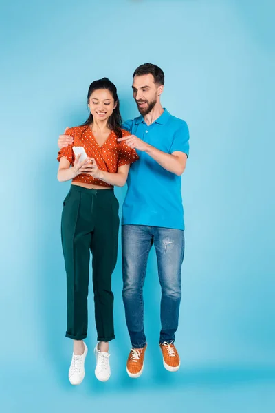 Barbudo hombre apuntando con el dedo en el teléfono inteligente en las manos de la mujer asiática mientras levita en azul - foto de stock