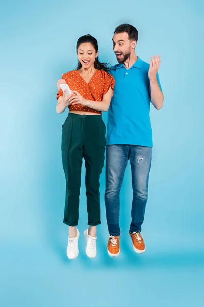 Excited man touching asian woman using smartphone while levitating on blue — Stock Photo
