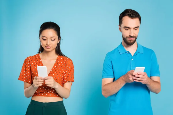Young asian woman in red blouse and bearded man in polo t-shirt using smartphones on blue — Stock Photo