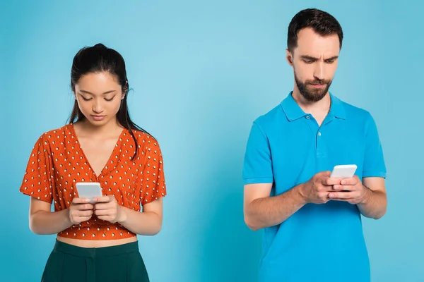 Morena mujer asiática en blusa roja y hombre en polo camiseta usando teléfonos inteligentes en azul - foto de stock