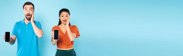 Website header of shocked interracial couple touching faces while showing smartphones with blank screen on blue — Stock Photo