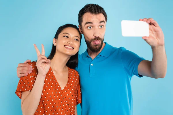 Asian woman showing peace sign near boyfriend sticking out tongue while taking selfie isolated on blue — Stock Photo