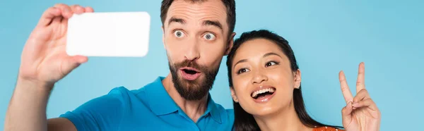 Panoramic concept of asian woman showing peace gesture near boyfriend grimacing while taking selfie isolated on blue — Stock Photo