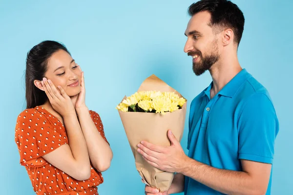 Young, bearded man in polo t-shirt presenting bouquet to pleased asian girlfriend in red blouse on blue — Stock Photo