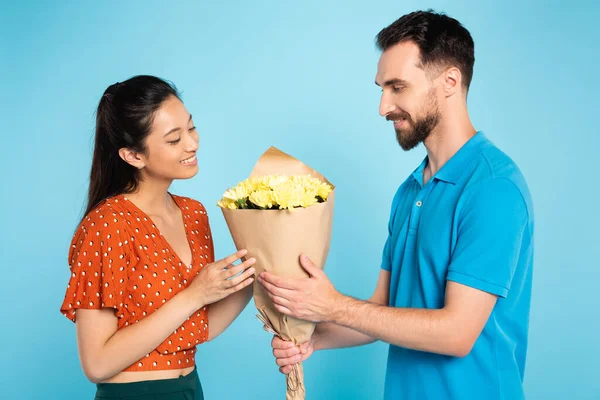 Giovane uomo in polo t-shirt presentando bouquet asiatico fidanzata in camicetta rossa su blu — Foto stock