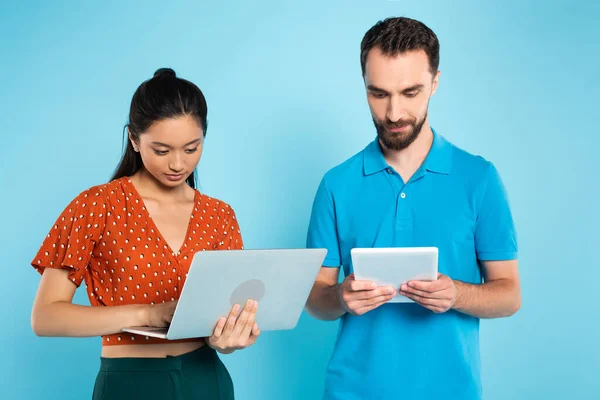 Young asian woman in red blouse using laptop near bearded man with digital tablet on blue — Stock Photo