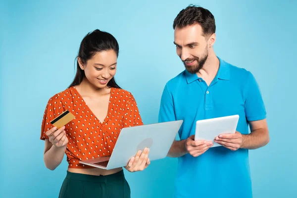 Brunette asian woman in red blouse holding credit card and laptop near bearded man with digital tablet on blue — Stock Photo