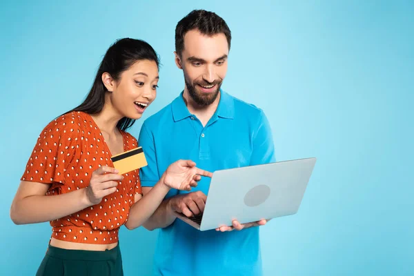 Joven hombre utilizando portátil cerca asiático mujer celebración de tarjeta de crédito y señalando con el dedo en azul - foto de stock