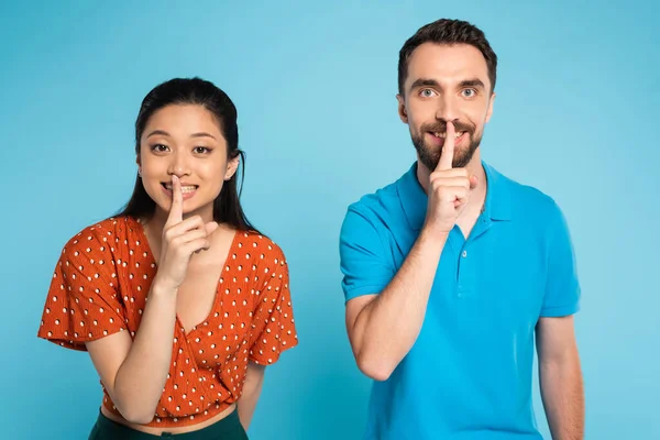 Joven interracial pareja mirando cámara mientras mostrando silencio gesto en azul - foto de stock