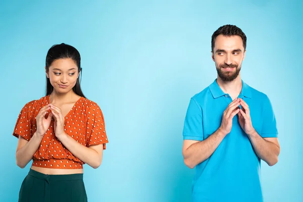 Femme asiatique sceptique en chemisier rouge et homme rusé en polo t-shirt geste avec les doigts joints sur bleu — Photo de stock