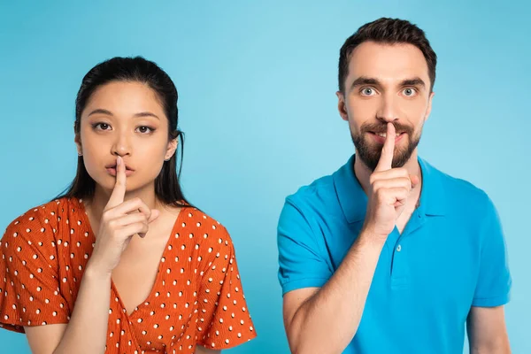 Jeune femme asiatique en chemisier rouge et homme en polo t-shirt montrant geste de silence sur bleu — Photo de stock