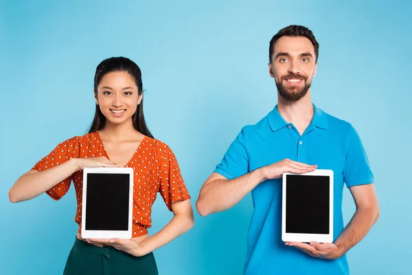 Азіатська жінка в червоній блузці і бородатий чоловік у футболці поло, що тримає цифрові планшети з порожнім екраном на синьому — стокове фото