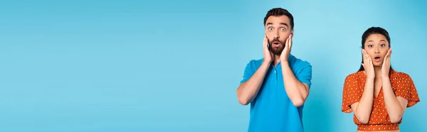 Choqué interracial couple toucher visages tout en regardant caméra avec des bouches ouvertes sur bleu, panoramique shot — Photo de stock