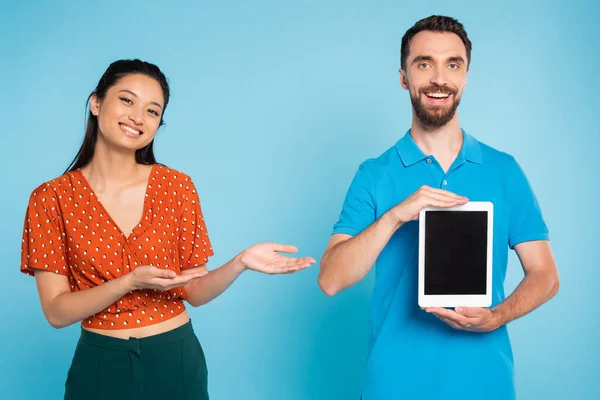 Barbudo homem no polo t-shirt mostrando digital tablet com branco tela perto asiático mulher apontando com as mãos no azul — Fotografia de Stock