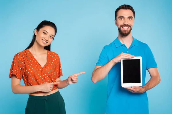 Brunette asian woman in red blouse pointing with fingers at digital tablet with blank screen in hands of bearded man on blue — Stock Photo