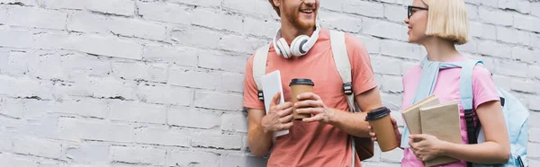 Panoramic crop of students holding paper cups and books near brick wall — Stock Photo