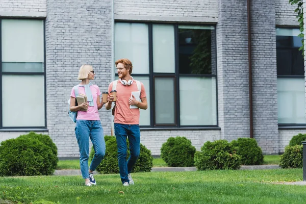 Students holding paper cups, books and digital tablet while walking on grass near campus — Stock Photo