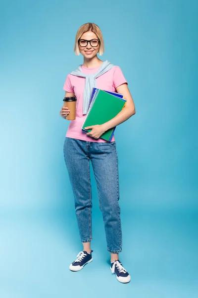 Estudante loira segurando copo de papel e cadernos enquanto está de pé em azul — Fotografia de Stock