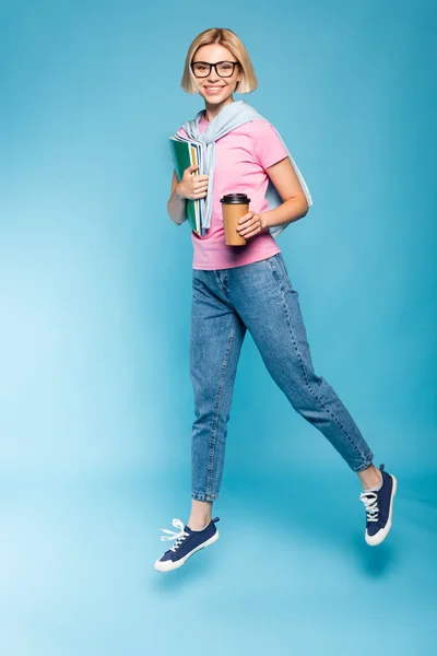 Blonde Studentin hält Pappbecher, Notizbücher in der Hand und fliegt auf blau — Stockfoto