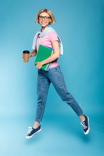 Young blonde student holding paper cup and notebooks while flying on blue — Stock Photo